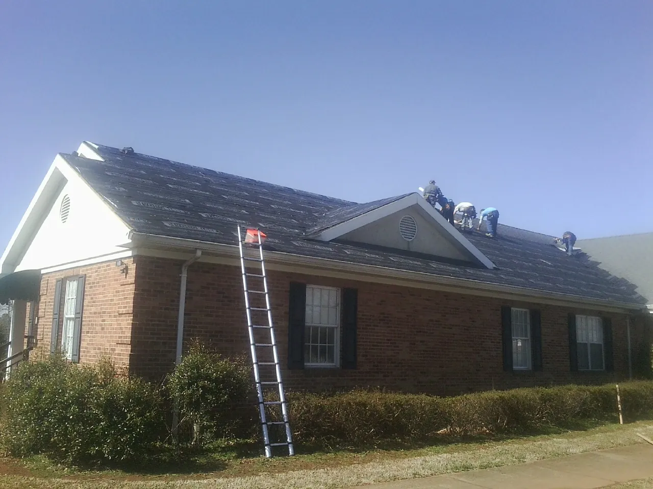 Crew tearing shingles off a brick ranch roof with a ladder set — Emergency Roof Repair work by our crew in La Harpe