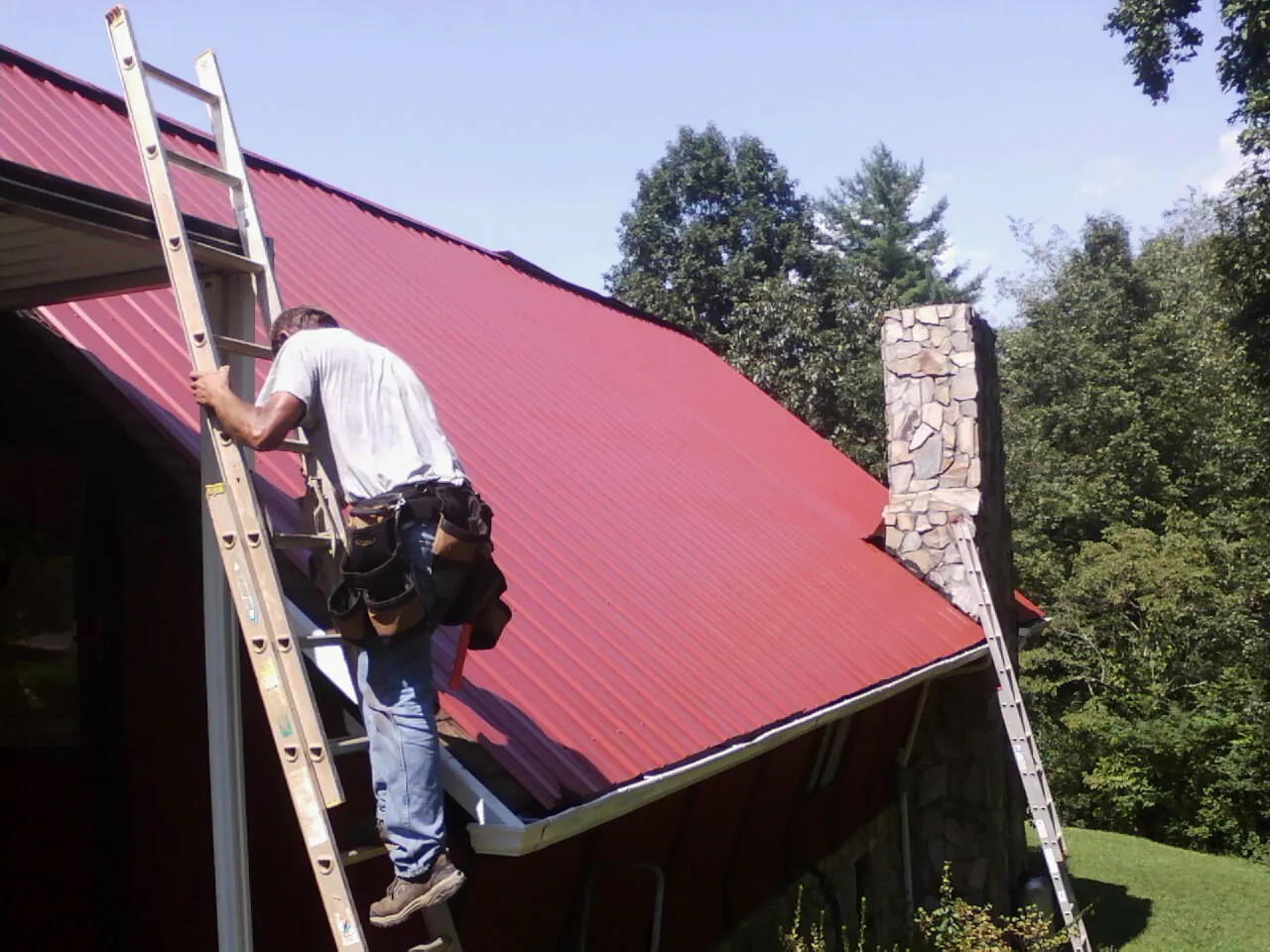 Roofer on a ladder beside a red metal roof and stone chimney — Soffit & Fascia Repair by Roofing Brothers in La Harpe